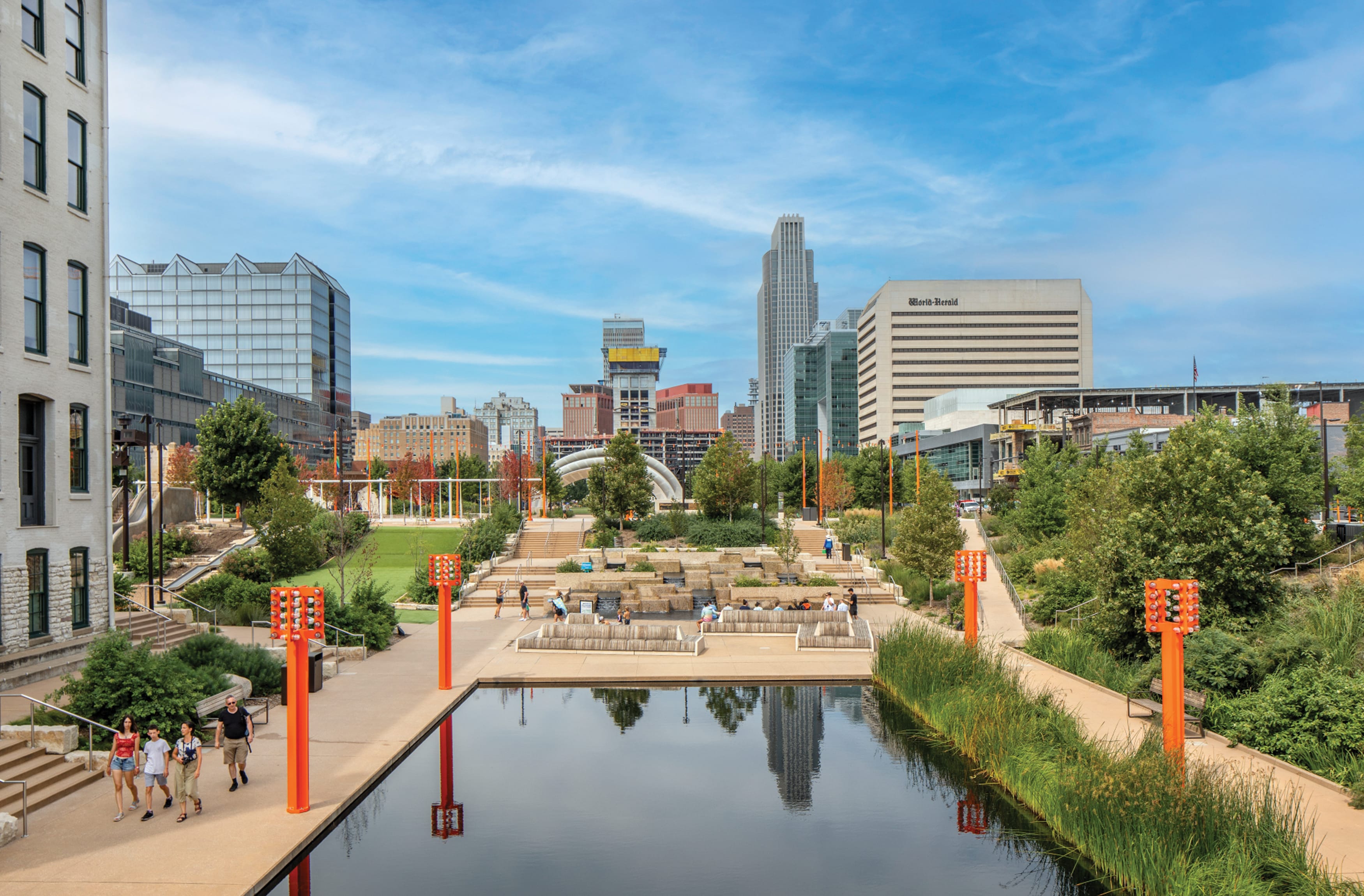 Pavilion with a manmade lake surrounded by native grass landscaping, custom sculptural concrete benches, and colorful industrial beam lampposts.